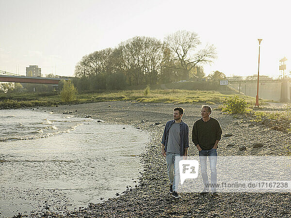 Son and father looking away while walking at riverbank