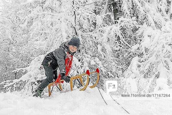 Boy sledding while playing with wooden sled on snow in forest