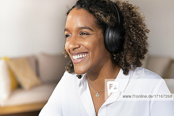 Fröhliche Frau mit Kopfhörern  die wegschaut  während sie zu Hause sitzt