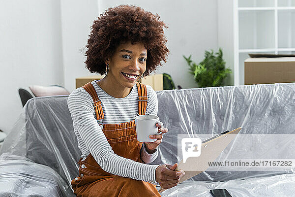 Lächelnde Frau trinkt Kaffee und hält eine Checkliste auf einem Klemmbrett in ihrem neuen Zuhause