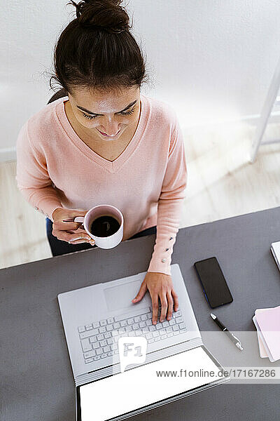 Woman with coffee cup working on laptop while sitting at home