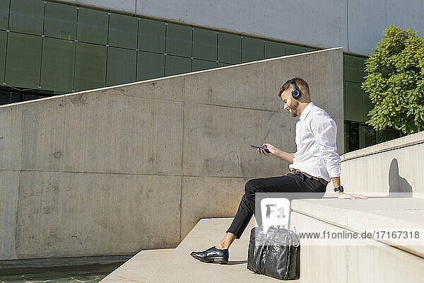 Businessman wearing headphones using mobile phone while sitting by briefcase on steps