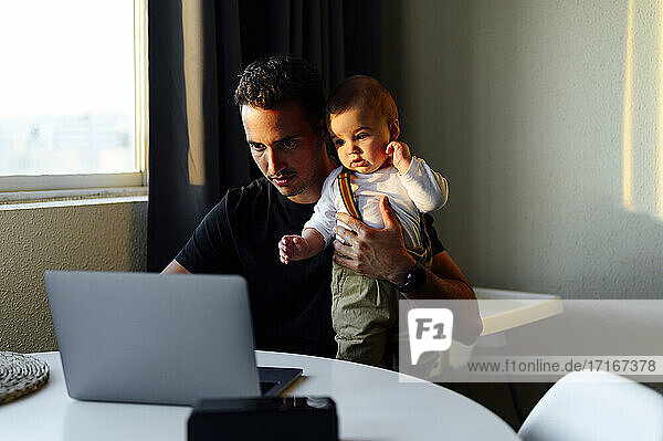 Father holding baby boy while working on laptop at home