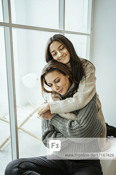 Mutter und Tochter umarmen sich mit geschlossenen Augen  während sie zu Hause auf der Fensterbank sitzen