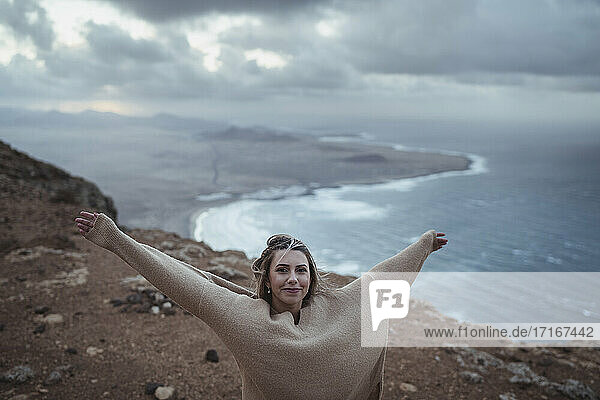 Lächelnde Frau  die mit ausgestreckten Armen auf einem Berg vor dem Strand von Famara steht  Lanzarote  Spanien