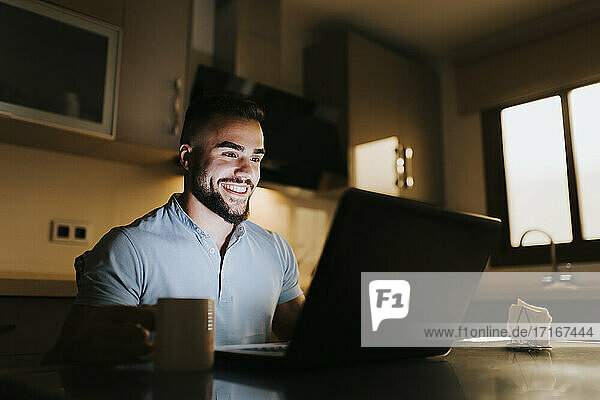 Lächelnder männlicher Unternehmer  der an einem Laptop arbeitet  während er am Esstisch in der Küche sitzt