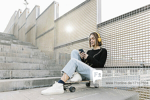 Junge Frau sitzt auf einem Skateboard und hört Musik im Freien