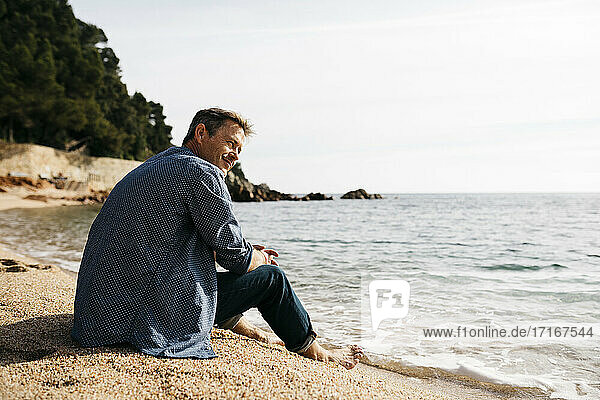 Lächelnder Mann am Strand sitzend und gegen das Meer blickend am Strand