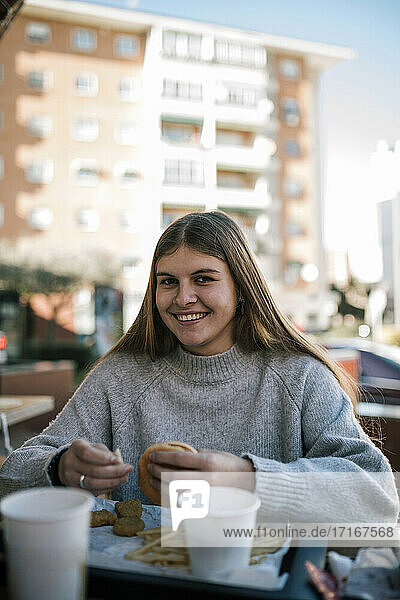 Lächelndes Teenager-Mädchen  das einen Burger isst  während es in einem Straßencafé sitzt