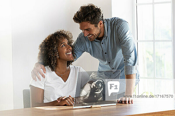 Smiling couple looking at each other while checking ultrasound image over transparent screen at home