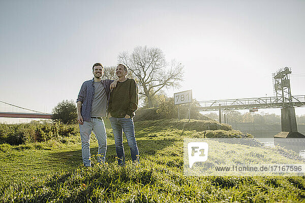 Smiling son and father standing on grass against clear sky at riverbank