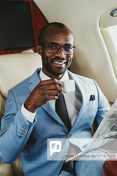 Glücklicher Geschäftsmann mit Kaffeetasse und Zeitung im Flugzeug