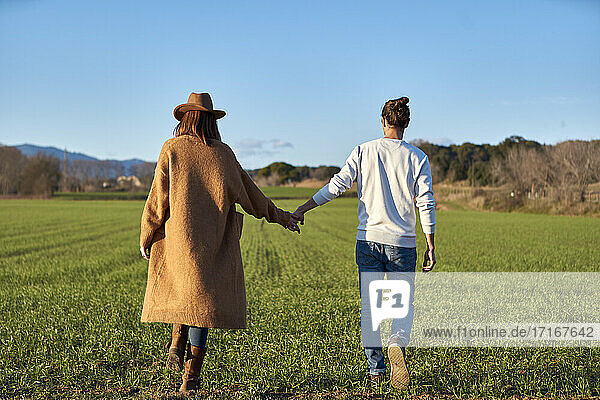Heterosexual couple holding hands while walking on green field during sunset