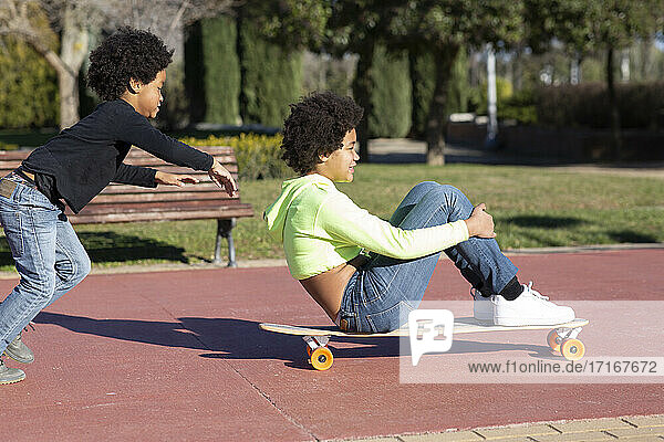 Bruder läuft hinter seiner Schwester  die auf einem Skateboard sitzt  während er im Park spielt