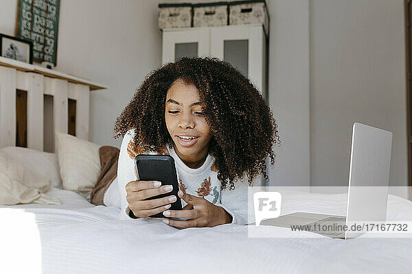Lockenköpfige Frau mit Laptop  die ein Mobiltelefon benutzt  während sie zu Hause auf dem Bett liegt