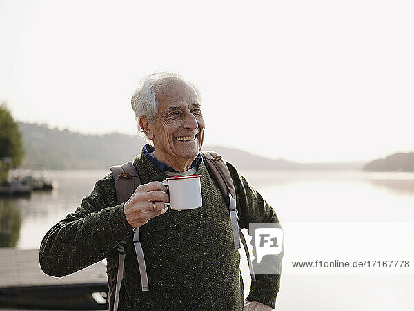 Lächelnder Mann mit Rucksack trinkt Kaffee  während er gegen den Himmel steht