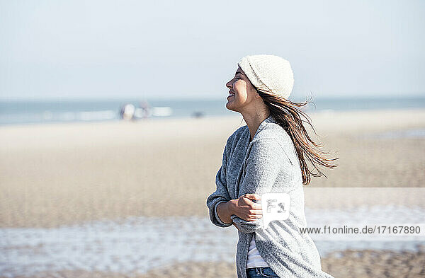 Glückliche junge Frau mit Strickmütze  die sich selbst umarmt  während sie am Strand steht