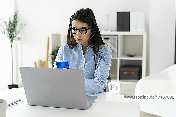 Berufstätige Frau  die ein Mobiltelefon benutzt  während sie mit einem Laptop am Schreibtisch im Büro sitzt