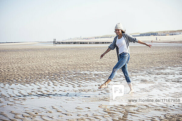 Unbeschwerte junge Frau mit Strickmütze  die auf nassem Sand am Strand spazieren geht