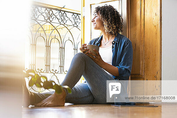 Young woman holding coffee mug while looking out of window at home