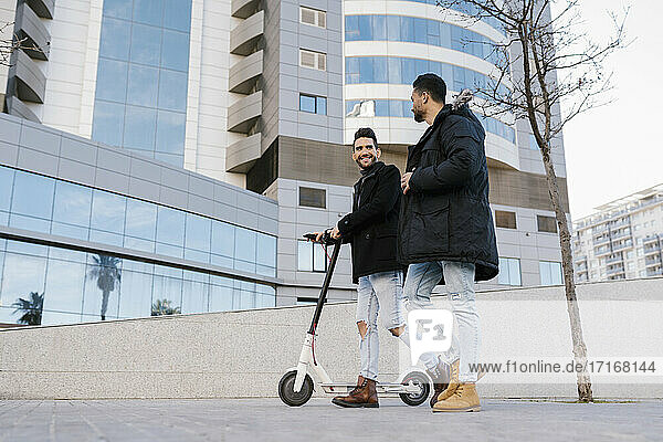 Mid adult man with electric scooter smiling at male friend while walking through city