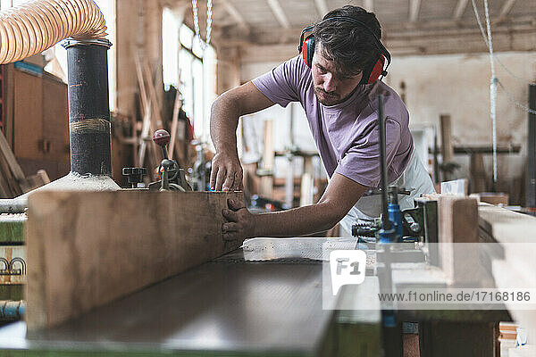 Young male craftsperson holding wooden plank on workbench while working in workshop