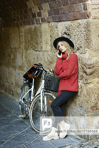 Young woman wearing hat talking on mobile phone while standing against wall by bicycle