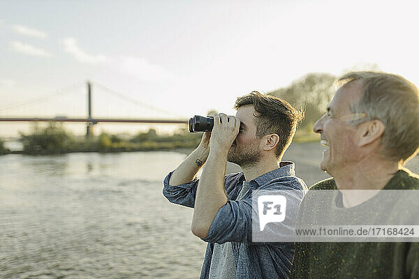 Son looking through binoculars while father laughing by on riverbank at evening