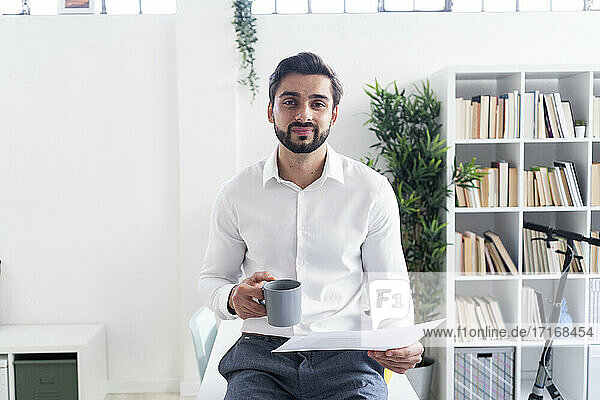 Lächelnde männliche Fachkraft mit Kaffeetasse hält Papierdokument am Arbeitsplatz