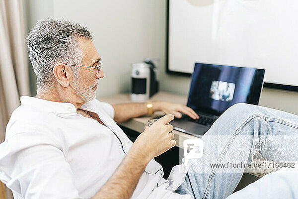 Mature man using laptop with feet up at desk while having coffee in hotel room