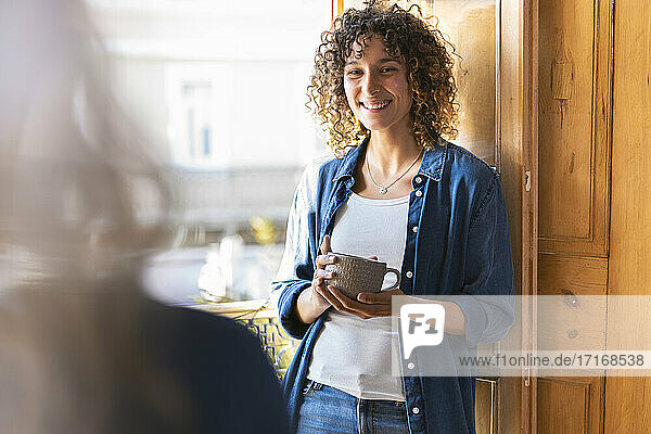 Happy curly woman looking at female friend while holding coffee mug