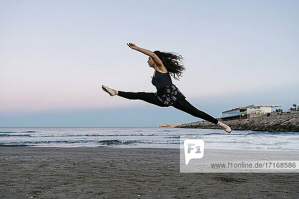 Graceful female ballet dancer with legs apart practicing at beach during sunset