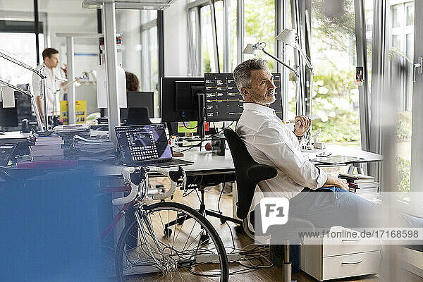 Geschäftsmann schaut weg  während er auf einem Stuhl mit Kollegen im Hintergrund in einem Großraumbüro sitzt