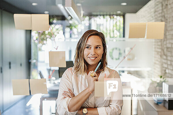 Lächelnde Geschäftsfrau mit Blick auf Haftnotizen  die an einer Glaswand im Büro kleben