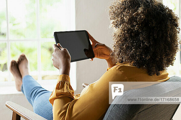 Frau mit lockigem Haar  die ein digitales Tablet benutzt  während sie sich auf einem Sessel zu Hause ausruht