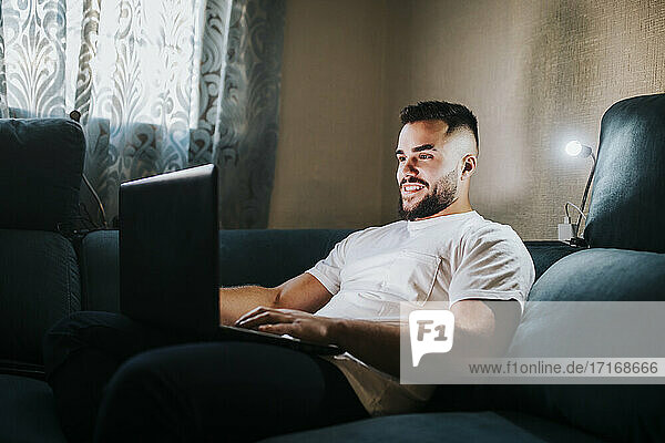 Lächelnder männlicher Unternehmer  der an einem Laptop arbeitet  während er auf der Couch im Wohnzimmer sitzt