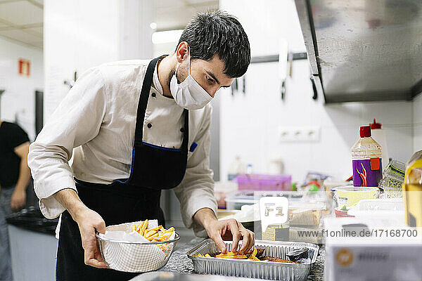 Männlicher Koch mit Schutzmaske ordnet Pommes frites in einem Folienbehälter in einer Restaurantküche an