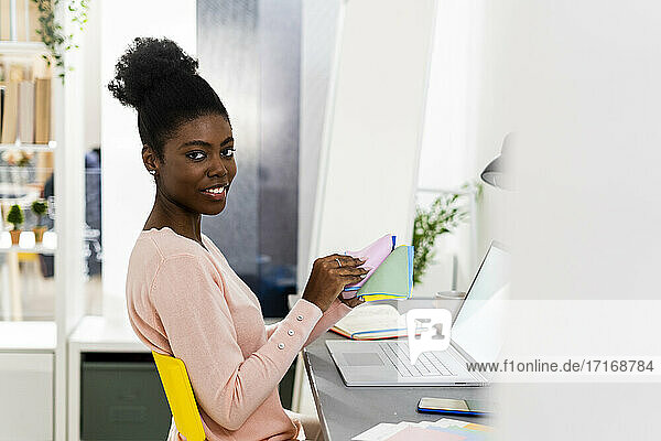 Lächelnde Geschäftsfrau  die ein buntes Stoffmuster hält  während sie zu Hause im Büro sitzt