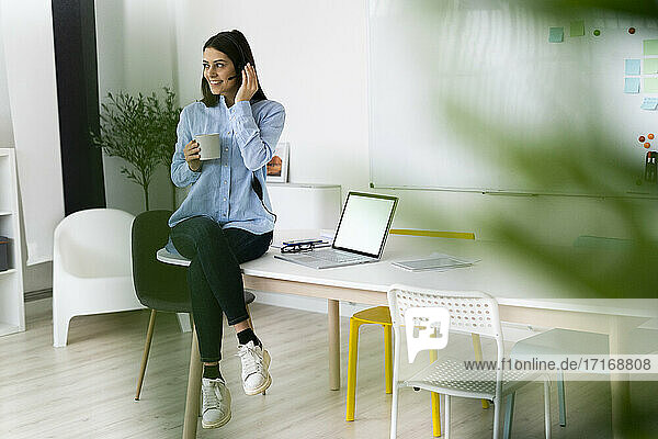Smiling businesswoman wearing headset drinking coffee while sitting on desk by laptop at office