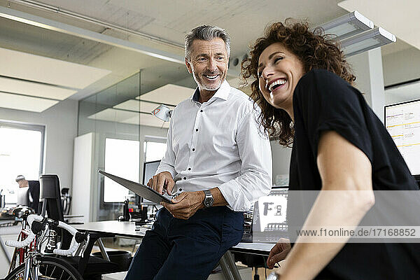 Lächelnder Geschäftsmann mit digitalem Tablet  der einen fröhlichen Kollegen ansieht  während er in einem Großraumbüro steht