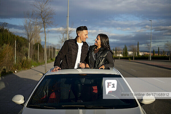 Junges Paar sieht sich an  während es auf dem Sonnendach eines Autos steht