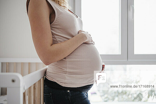 Pregnant woman with hands on stomach standing by window at home