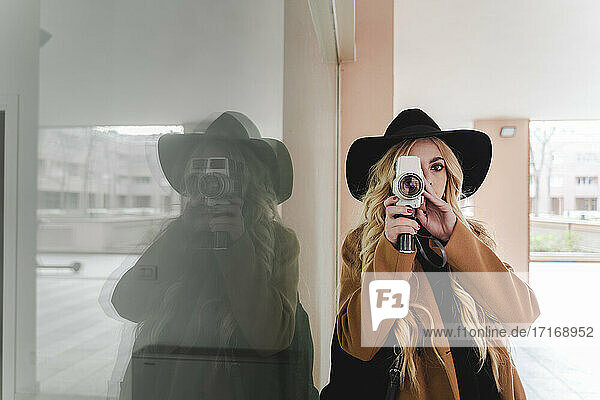 Young woman in hat filming through vintage video camera against reflection in window