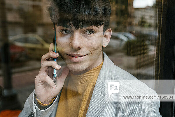Lächelnder Mann  der durch das Fenster schaut  während er in einem Café mit seinem Mobiltelefon spricht