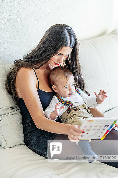 Young mother showing book to baby boy while sitting on bed at home
