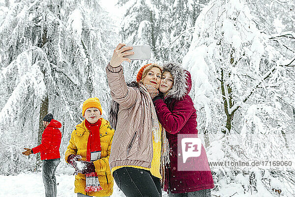 Mother and daughter taking selfie through mobile phone while standing with family in background at forest