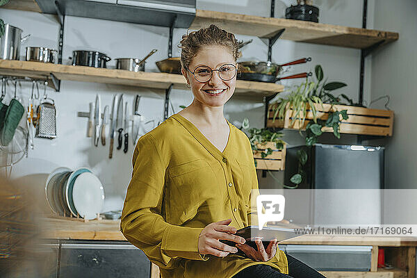 Young woman wearing eyeglasses smiling while using digital tablet in kitchen