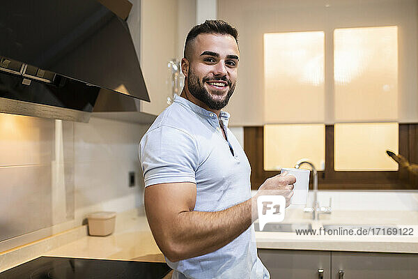 Muscular young man smiling while holding coffee cup in kitchen