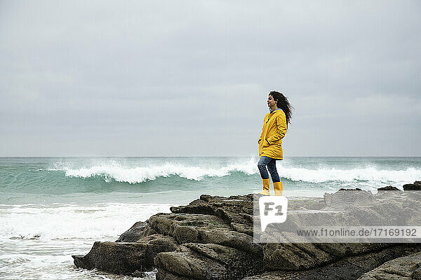 Junge Frau in gelbem Regenmantel  die auf einem Felsen stehend das Meer betrachtet