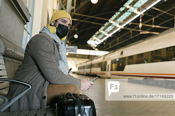 Mann mit Gesichtsschutz  der auf einer Bank im Bahnhof sitzt und ein Mobiltelefon benutzt